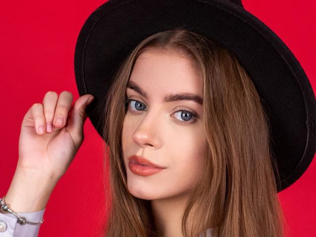 EricaDiaz with long brown hair, blue eyes, and light skin, wearing a black hat, holding it with her right hand, and a silver bracelet, against a bright red background.