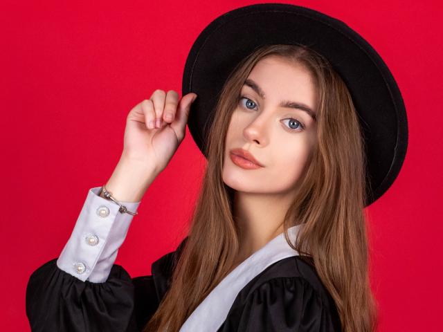 EricaDiaz with blue eyes, wearing a black hat and black-and-white dress, touching her hat with a silver bracelet on her wrist, against a red background.