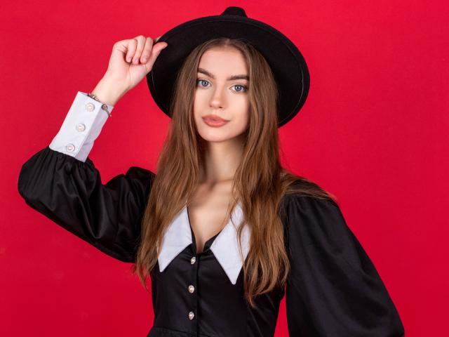 EricaDiaz with long brown hair, wearing a black hat, black shirt with white collar, and buttons, touching her hat against a red background. She's got a cute face.