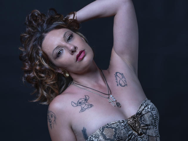 Cerisette with curly brown hair, red lipstick, tattoos on her tits and arms, snake-patterned dress, necklace, arm raised, dark background, looking at camera, slightly tilted head.
