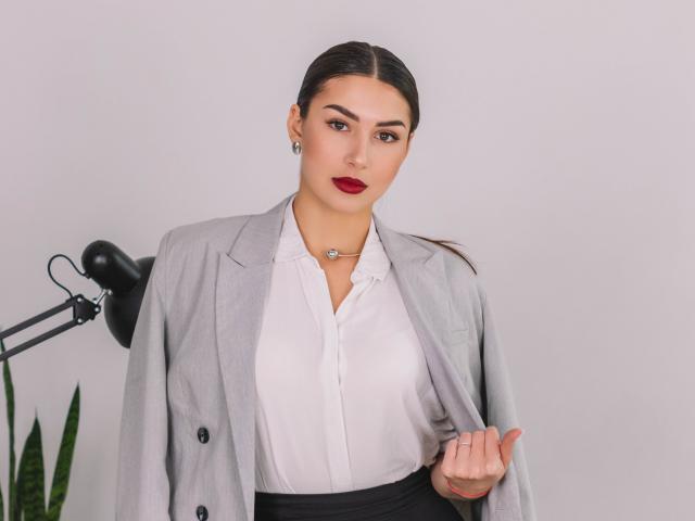 OliviaCluff has dark brown hair, red lipstick, and is wearing a gray blazer over a white button-up shirt, black skirt, and necklace, with a black lamp to the left.