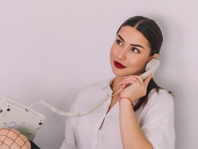 OliviaCluff with dark hair, red lipstick, white shirt, holding a corded phone to her ear, wearing fishnet stockings, looking to the side with a slight smile.