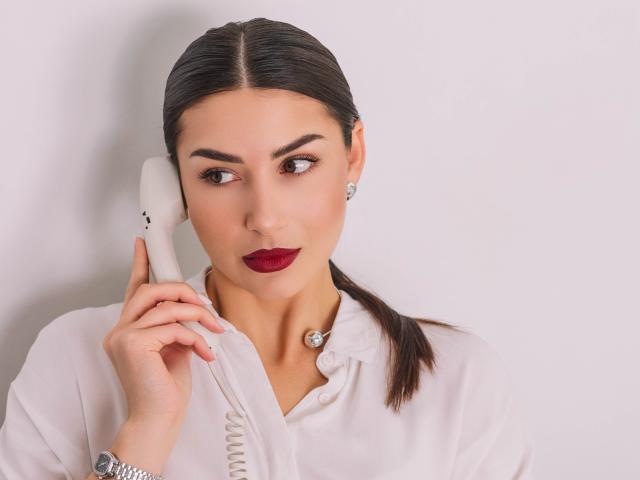 Olivia Cluff, met donkerbruin haar in een paardenstaart, houdt een witte, gesnoerde telefoon aan haar oor, draagt een witte blouse met knopen en rode lippenstift, en ziet er licht geïrriteerd uit..