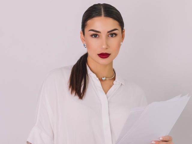OliviaCluff with dark hair in a ponytail, wearing a white button-up shirt, holding papers, red lipstick, pearl necklace, and earrings, looking at the camera.