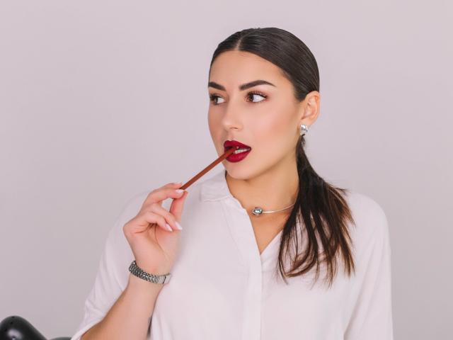 OliviaCluff with slicked-back brown hair, wearing a white button-up, red lipstick, holding a red lipstick tube to her mouth, silver bracelet, and diamond earrings.