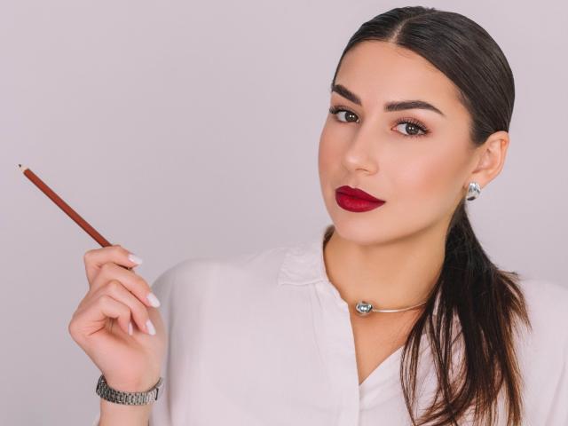 OliviaCluff with dark hair in a ponytail, red lipstick, white blouse, holding a brown pencil, wearing a silver necklace and earrings, and a silver bracelet.