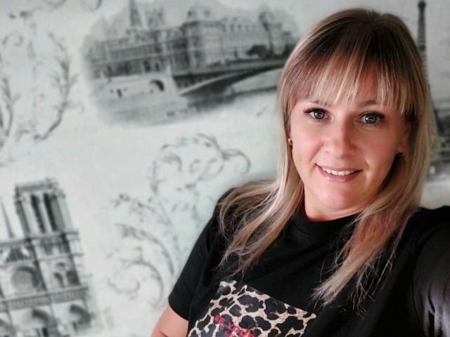 MirandaStone with blonde hair, bangs, and a black t-shirt with a leopard print design, smiling at the camera, showing her cute face and small, round tits.