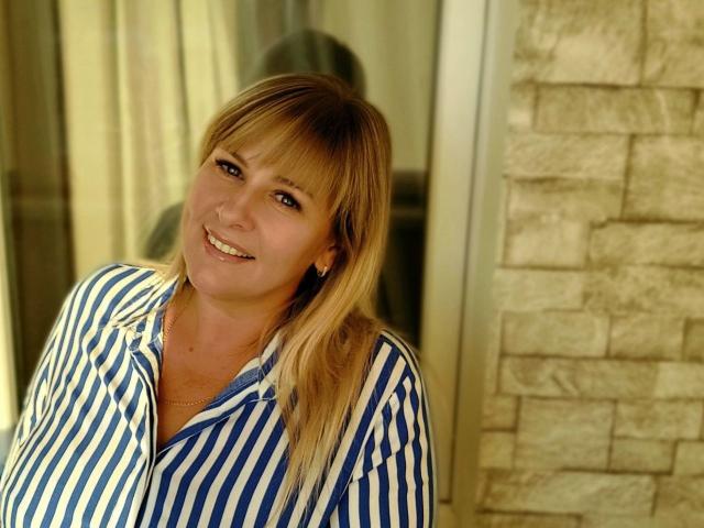 MirandaStone with blonde hair, blue and white striped shirt, smiling, has small tits, standing near a stone wall, reflection in glass behind her.
