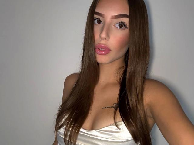 NicaDiamond-hot has long brown hair, full pink lips, and a tattoo above her tits. She's wearing a white strapless top, looking at the camera with a pouty expression.