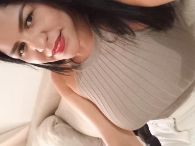 IsabellaTania with dark hair, red lipstick, and a beige ribbed sleeveless top, showing her tits, smiling at the camera, sitting on a white couch.