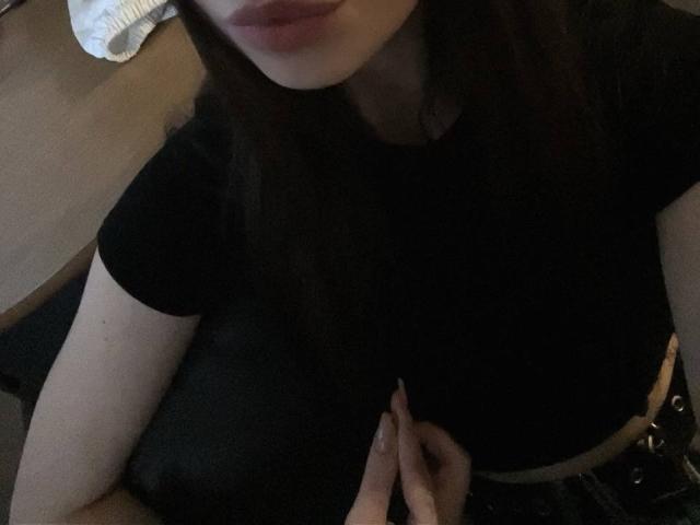 Amabel's face is partially visible, showing pink lips. She's wearing a black shirt, with her hand on her bare belly. Dark brown hair, pale skin, dim lighting.