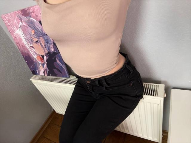 Amabel's pale skin and flat chest are visible in a tight, beige top. She's wearing black jeans, standing with her back to a white radiator. Anime poster in the background.
