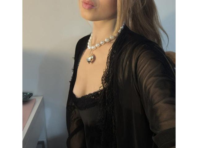 StellinaVelvet with puffy lips, wearing a black lace-trimmed top, sheer black cardigan, and white pearl necklace with a heart pendant, showing her tits. Brown hair, slightly wavy.