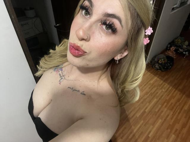 SammyViolet with blonde hair, pink flower clip, nose ring, and red lipstick, pouting, showing big tits, tattoo on chest, black strap dress, wooden floor, and eyelashes.