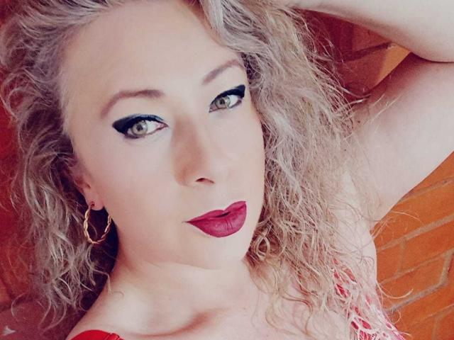 SofiaCurvyCute has curly blonde hair, green eyes with black eyeliner, red lipstick, and gold hoop earrings, lying with one hand in her hair. Her boobs are partially visible.
