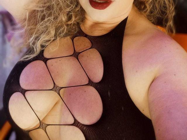 SofiaCurvyCute with curly blonde hair, wearing a black mesh top, exposing her large tits and pinkish areolas, with dark red lipstick.