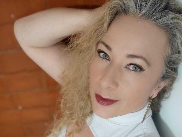 SofiaCurvyCute has curly blonde hair, green eyes, and red lipstick. She's wearing a white top, with one arm raised, showing off her tits. She's smiling subtly.