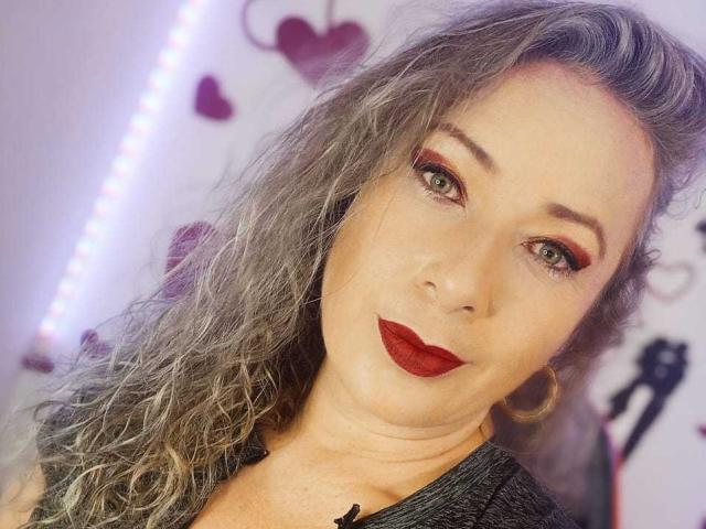 SofiaCurvyCute has curly gray hair, red eyeliner, green eyes, and red lipstick. She's wearing a black top and gold hoop earrings, smiling at the camera.
