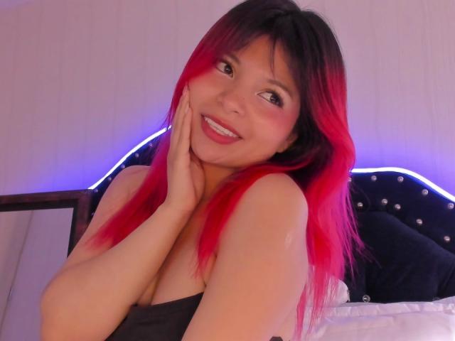 ShofiaWett with red and black hair, smiling, hand on cheek, showing cleavage in a black top, blue-lit headboard, and white pillow in the background.