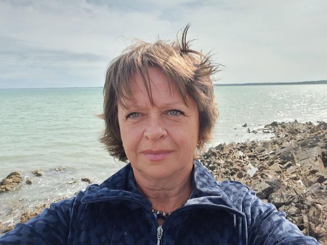 MaltaFora has short brown hair, green eyes, and a slight smile. She wears a blue zip-up hoodie and stands by a rocky shoreline with a calm sea.