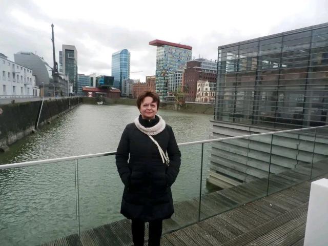 MaltaFora stands on a wooden bridge, wearing a black coat with a white scarf, hands in pockets, smiling. She has brown hair and is in front of a city waterway.