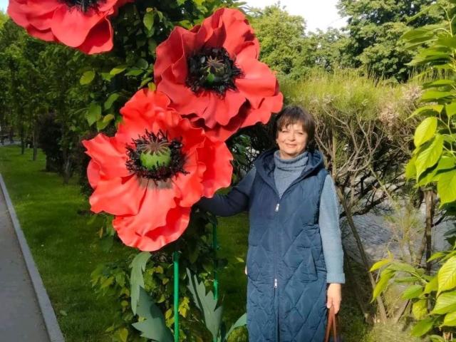 MaltaFora stands with a blue quilted coat, gray turtleneck, and brown bag, holding a tree with huge red poppy flowers. Green grass and trees in the background.