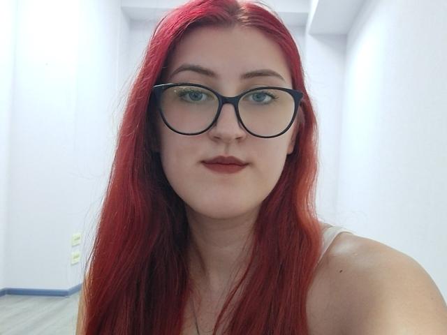 Triziya, met rood haar, draagt zwarte brillen, heeft groene ogen en rode lippenstift. Ze kijkt naar de camera met een lichte glimlach en draagt een witte tanktop..