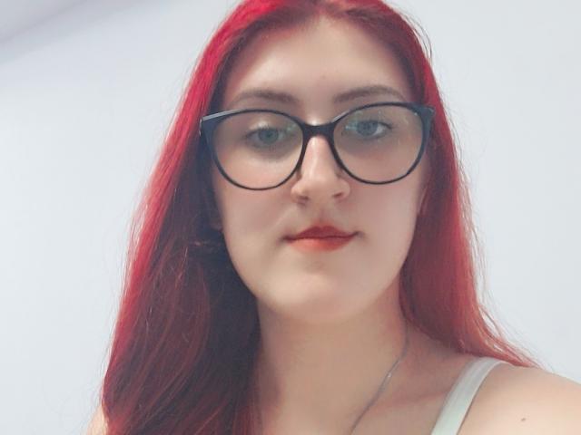 Triziya with red hair, wearing black glasses, white tank top, and red lipstick, showing her fair skin and subtle smile. Her tits are not visible, only her face.