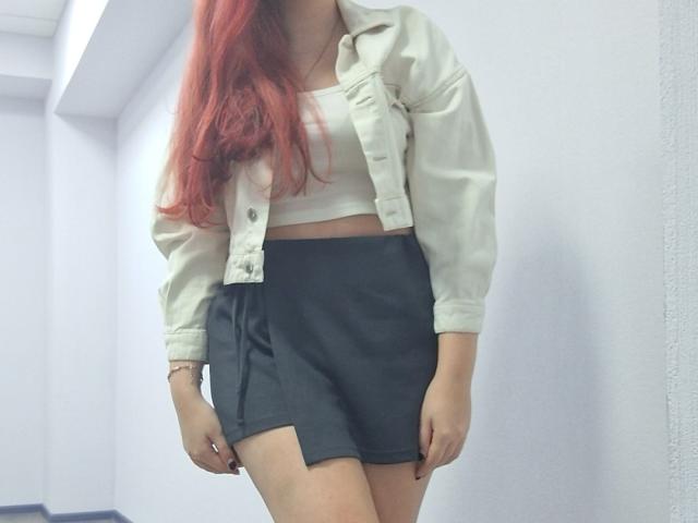 Triziya, met rood haar, draagt een witte blouse en een zwarte rok. Haar borsten zijn bedekt, maar ze laat een beetje van haar achterwerk zien, in een gang met witte muren..