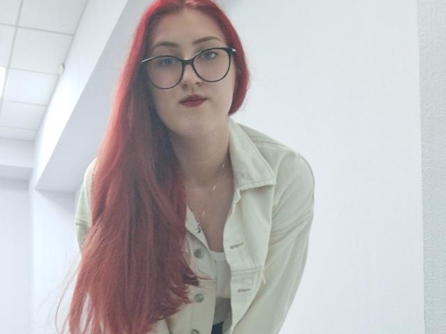 Triziya with long red hair, wearing glasses and a white button-up shirt, leaning forward, showing her tits, has red lipstick, and a subtle necklace.