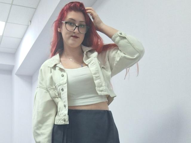 Triziya with red hair, glasses, white crop top, and black skirt, wearing an open white denim jacket, hand in hair, standing in a white-walled room.