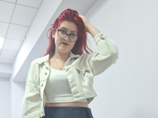 Triziya with red hair, glasses, white crop top, and beige jacket, hand in hair, smiling, showing her ass, standing in a white-walled room.
