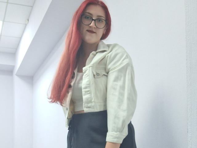 Triziya with long red hair, wearing glasses, a white denim jacket, and black pants, standing in a white hallway, showing a confident, slightly smug expression.