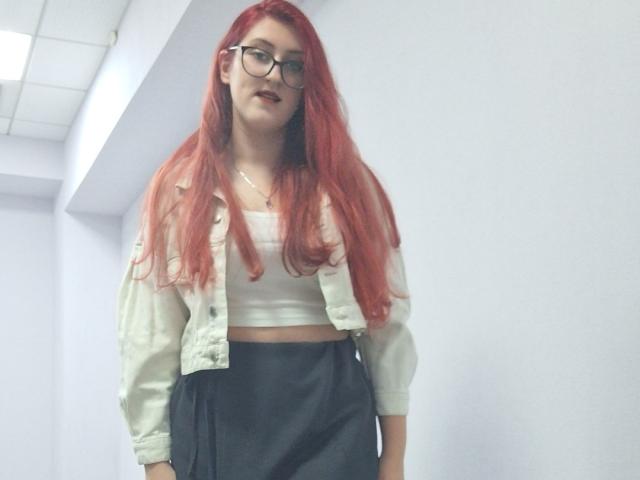 Triziya with long red hair, glasses, white crop top, green jacket, and black skirt, standing in a white hallway. Boobs are small, face is cute, and she looks confident.