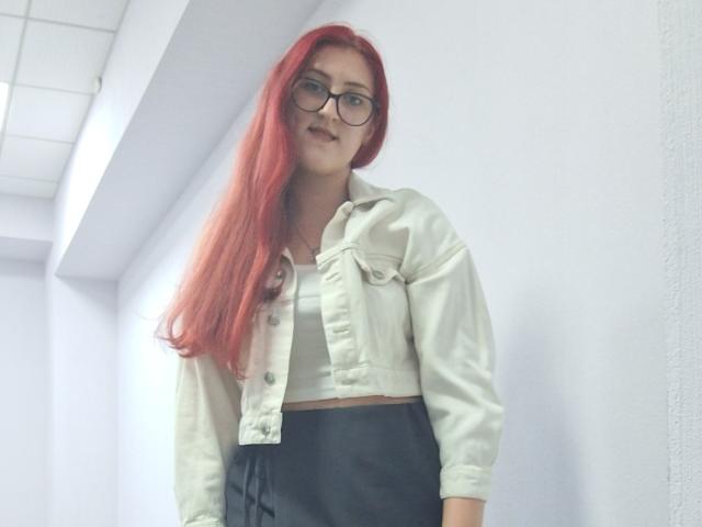 Triziya with long red hair, glasses, white shirt jacket, black skirt, standing in a white-walled room, looking slightly down. Her tits are small, and she has a cute face.