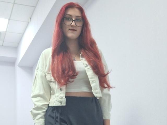 Triziya with long red hair, wearing glasses, a white crop top, and black high-waisted pants, and a beige jacket. Her tits are small and she's standing in a hallway.