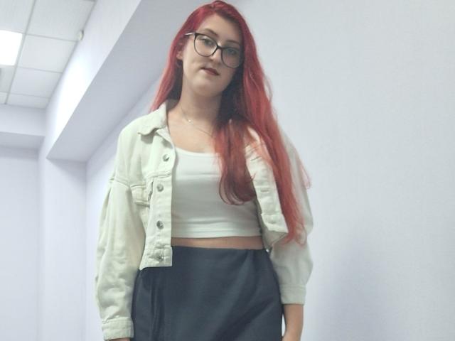 Triziya with red hair, glasses, white crop top, and black skirt, wearing a light green jacket, standing in a white-walled hallway. Her ass is covered by the skirt.