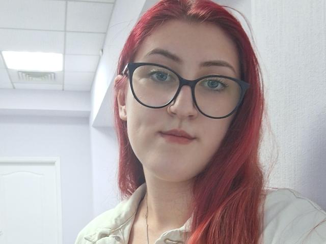 Triziya with red hair, glasses, and a slight smile, wearing a green shirt, has fair skin and a cute, nerdy look. Her tits are small and she's attractive.
