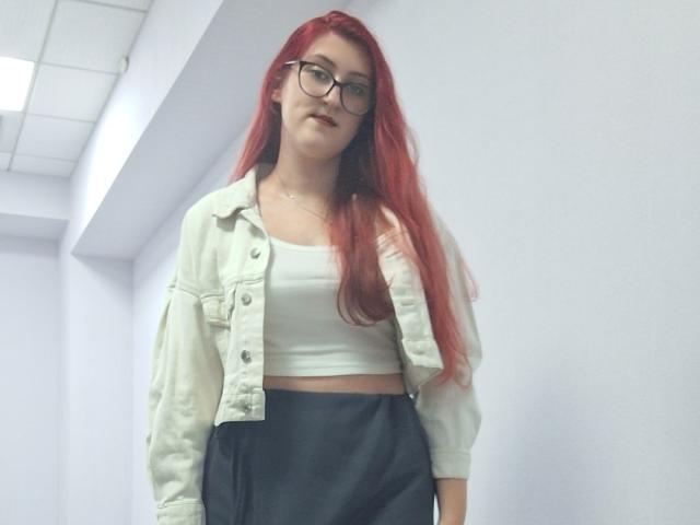 Triziya with red hair, glasses, white crop top, and black high-waisted pants, wearing a light green jacket, standing in a white hallway. Her tits are small.