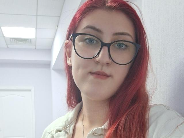 Triziya with red hair, black glasses, and pale skin, wearing a beige shirt, has a neutral expression, and is in a brightly lit room.