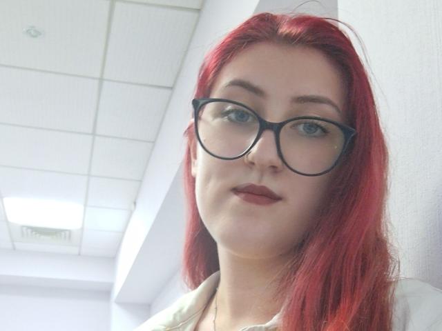 Triziya with red hair, black glasses, and light skin, showing her face, has small tits, a cute smile, and wears a white shirt, in a bright room.
