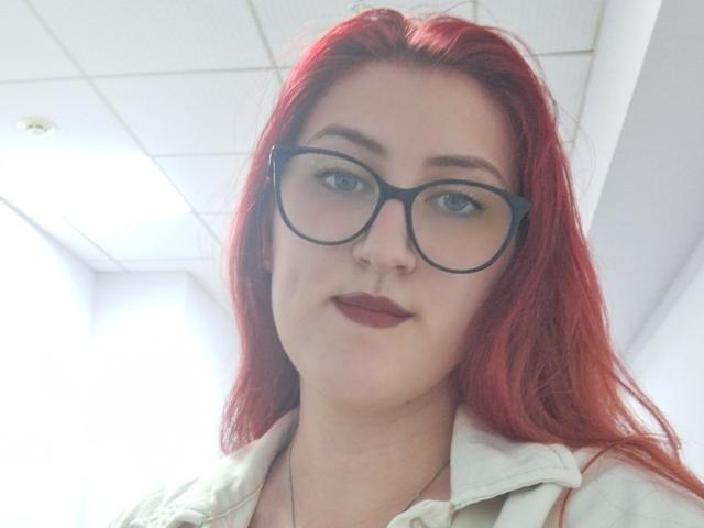 Triziya with red hair, glasses, and dark lipstick, wearing a white shirt, has a slight smile and fair skin, photographed from a low angle.