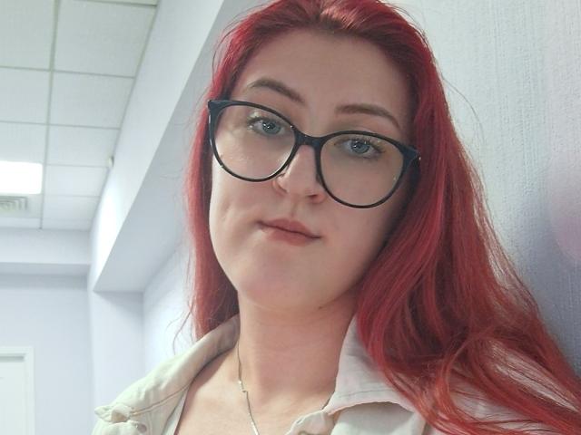 Triziya with red hair, black glasses, light skin, and subtle smile, wearing a beige shirt, has small tits and a silver necklace, in a white-walled room.