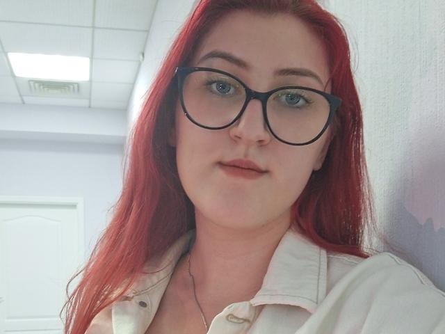 Triziya with red hair, black glasses, and light skin, wearing a white button-up shirt, showing a subtle smile, standing against a white wall.