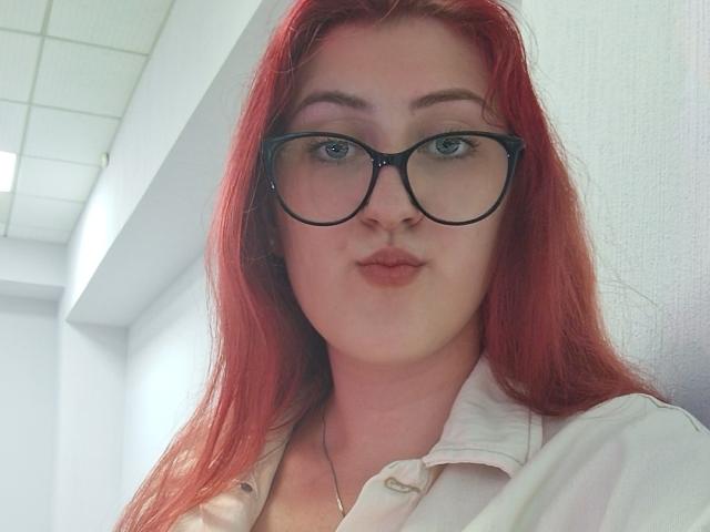 Triziya has red hair, glasses, and is pouting with pink lips. She's wearing a white shirt, and her blue eyes are looking directly at the camera.