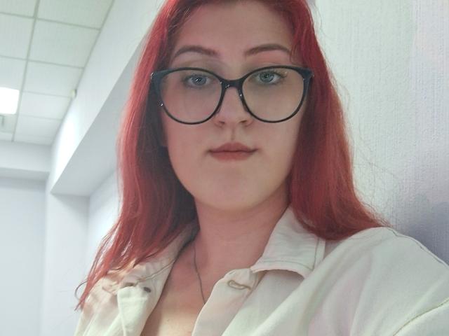 Triziya has red hair, black glasses, and is wearing a white button-up shirt. She's looking at the camera with a slight smile. Her tits are visible under the shirt.