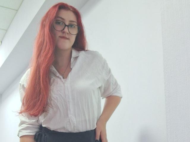 Triziya with long red hair, glasses, white button-up shirt, black skirt, standing, hands on hips, slight smile, white wall, ceiling light visible.
