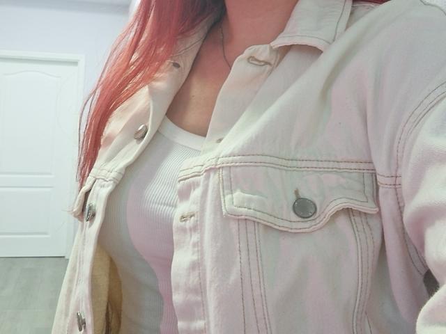 Triziya's pinkish jacket over a white ribbed shirt, showing her small tits, with long red hair partially visible, in a bright room.