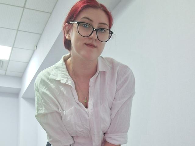 Triziya with red hair, glasses, and a white button-up shirt, leaning forward, showing her tits slightly through the shirt. She's smiling subtly, looking cute.