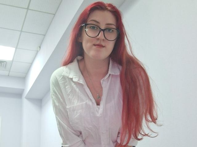 Triziya with long red hair, wearing glasses and a white button-up shirt, has a cute smile and small tits, standing in a white hallway.